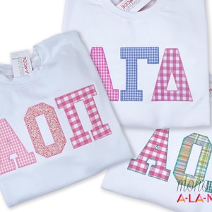 May include: White t-shirts with embroidered Greek letters in various patterns, including gingham and floral prints. The shirts feature the letters "AOT" and "ITA". The brand name "monogram A-LA-MODE" is visible.
