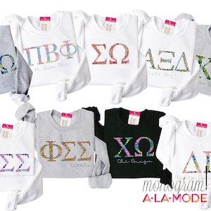 Könnte beinhalten: Eine Sammlung von acht langärmeligen Sweatshirts mit floralen Buchstaben-Designs, die verschiedene Sororitäten repräsentieren. Die Sweatshirts sind in verschiedenen Farben, darunter Weiß, Grau und Schwarz. Die Sorority-Buchstaben sind vollständig ausgeschrieben, darunter "Chi Omega", "Delta Gamma", "Phi Sigma Sigma", "Sigma Sigma Sigma", "Alpha Delta Kappa", "Sigma Omega", "Pi Beta Phi" und "Zeta Sigma Alpha".