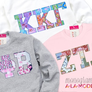 May include: Three sweatshirts with sorority letters in patchwork fabric. The letters are KKI, IΦB, and ZTA. The sweatshirts are white, gray, and pink. The text "monogram A-LA-MODE" is at the bottom of the pink sweatshirt.