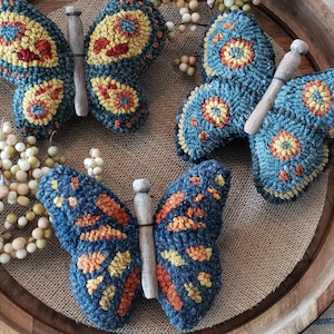 Pode incluir: Tr&ecirc;s ornamentos de borboleta de feltro azul e laranja com pinos de madeira. As borboletas est&atilde;o dispostas em uma bandeja de madeira marrom com tecido de juta.