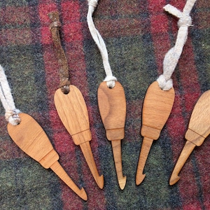 May include: Five wooden crochet hooks, each featuring a smooth, light brown finish and a distinct handle shape. Each hook is attached to a short, knotted fabric cord in varying colors, including beige, brown, and green, for hanging or storage purposes.