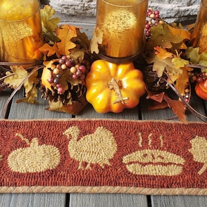 May include: A brown and tan wool rug runner with a Thanksgiving theme. The rug has five designs: a pilgrim hat, a pumpkin, a turkey, a pie, and a leaf. The rug is on a wooden table with a fall centerpiece of leaves, gourds, and candles.