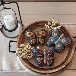 Pode incluir: Tr&ecirc;s ornamentos de ponto de agulha em forma de borboleta azul e amarela em uma bandeja de madeira com bagas brancas.