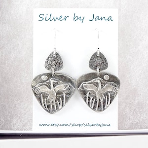 May include: Silver earrings with a textured design featuring a pair of birds in flight. The earrings are silver and have a textured finish. The earrings are hanging from a silver hook.