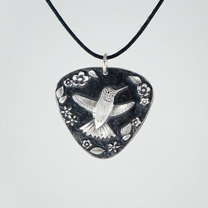 May include: A silver pendant necklace with a black cord. The pendant is shaped like a guitar pick and features an engraved hummingbird with floral details.