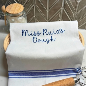 May include: White kitchen towel with blue stripes and the embroidered text "Miss Ruiz's Dough". The towel is folded over a woven basket. A jar with a wooden lid and a dough whisk are also visible.