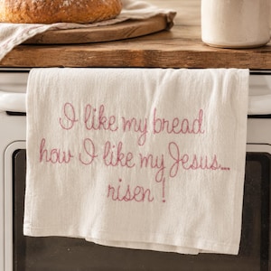 Puede incluir: Un paño de cocina blanco con texto bordado en rosa: "I like my bread how I like my Jesus... risen!" En el fondo se ven un pan y una taza. El pan está sobre una tabla de cortar.