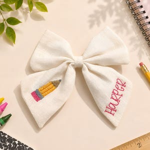 May include: An off-white fabric hair bow featuring an embroidered yellow pencil on the left tail and the name Harper embroidered in pink thread on the right tail. The bow is positioned near a wooden ruler showing centimetres and school supplies.
