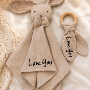 Puede incluir: Un arrullo para beb&eacute; con forma de conejito beige y un mordedor de madera. El arrullo tiene una pajarita y el texto bordado "Love You". El mordedor tambi&eacute;n tiene el texto "Love You". Ambos art&iacute;culos son de tela suave y texturizada.
