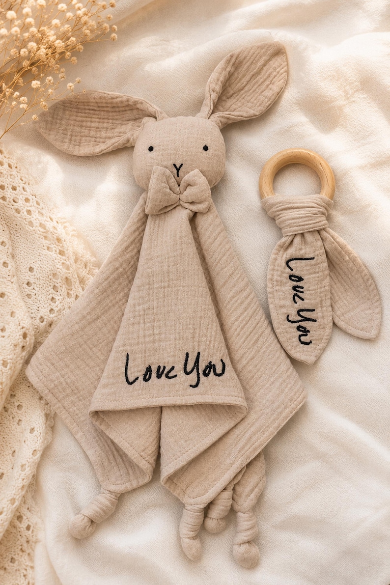 Puede incluir: Un arrullo para beb&eacute; con forma de conejito beige y un mordedor de madera. El arrullo tiene una pajarita y el texto bordado "Love You". El mordedor tambi&eacute;n tiene el texto "Love You". Ambos art&iacute;culos son de tela suave y texturizada.
