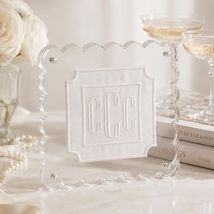May include: A square clear acrylic frame with a scalloped edge featuring a white fabric insert in the center. The fabric is embroidered with the white monogram letters CCC in a classic serif font, secured by four small silver metal corner bolts.