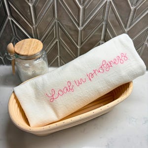 Puede incluir: Un paño de lino blanco con la frase "Loaf in progress" bordada en rosa, dentro de una cesta de mimbre. Un tarro de cristal con tapa y cuchara de madera, lleno de harina, está a la vista.