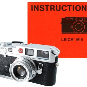 May include: A silver Leica M6 film camera with a black leather covering and a black lens with the text "Leitz Elmar 1:2.8 15cm Nr. 1602701" printed on it. The camera is next to a red instruction manual with the text "INSTRUCTIONS" and "LEICA® M 6" printed on it.