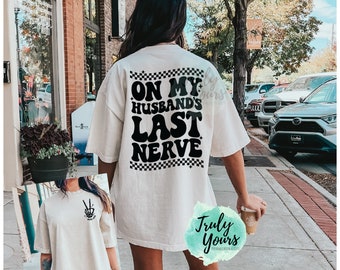 May include: An off-white short-sleeve t-shirt featuring black text on the back that reads ON MY HUSBAND'S LAST NERVE with checkered borders. The front of the shirt displays a small black graphic of a hand making a peace sign with the text WIFE LIFE.