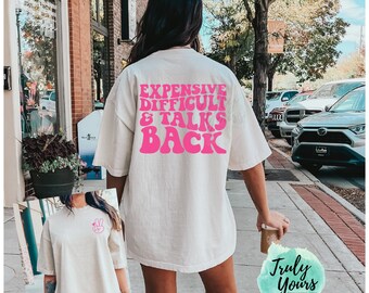 May include: An off-white short-sleeve t-shirt featuring bright pink text on the back that reads EXPENSIVE DIFFICULT & TALKS BACK in a bold, wavy font. The front of the shirt displays a small pink peace sign graphic with a heart on the left chest.