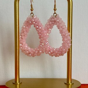 Könnte beinhalten: Ein Paar rosa Tropfenohrringe aus Perlen. Die Ohrringe bestehen aus kleinen, hellrosa Perlen und haben goldfarbene Ohrhaken. Die Ohrringe werden auf einem goldfarbenen Ohrringständer vor einem weißen Hintergrund präsentiert.