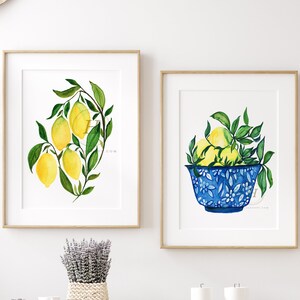 May include: Two framed watercolor paintings of lemons. The first painting shows a branch of lemons with green leaves. The second painting shows a blue and white bowl filled with lemons and green leaves.