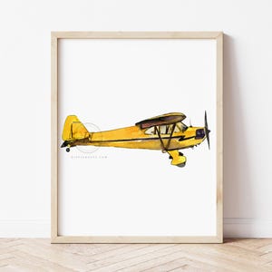 Puede incluir: Impresión en acuarela de un avión amarillo en un marco de madera. El avión tiene una parte superior marrón y una hélice. La obra de arte está sobre un fondo blanco y se exhibe sobre un suelo de madera clara.