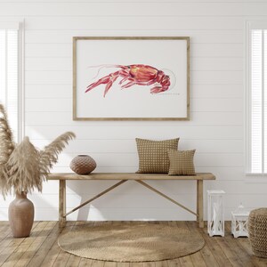 May include: A watercolor painting of a red crayfish on a white background. The painting is framed in a light wood frame and is hanging on a white wall above a wooden bench.
