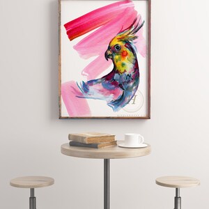 May include: A framed watercolour print of a colourful cockatiel with a yellow crest and pink brushstrokes. The artwork is displayed on a white wall above a small round table with books and a teacup.