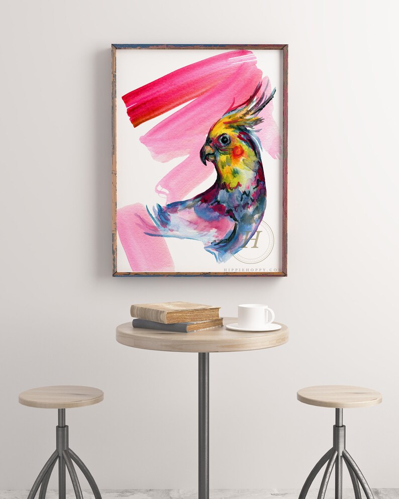 May include: A framed watercolour print of a colourful cockatiel with a yellow crest and pink brushstrokes. The artwork is displayed on a white wall above a small round table with books and a teacup.
