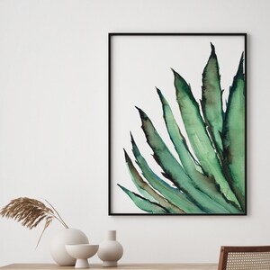 May include: Watercolor painting of a green agave plant in a black frame. The artwork features long, pointed leaves with dark green and blue-green hues against a white background. The painting is displayed on a wall above a wooden table with decorative vases.
