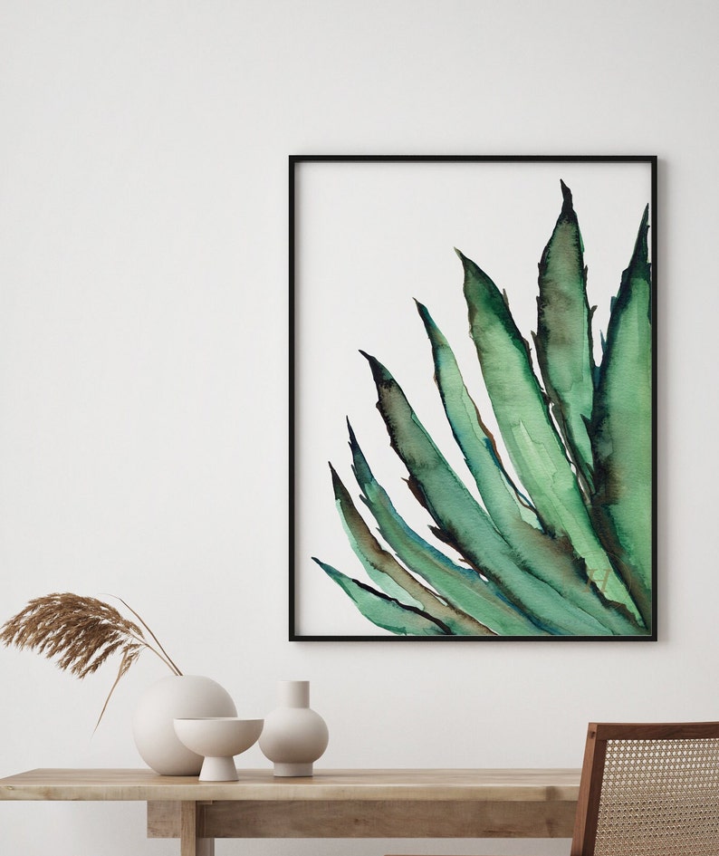May include: Watercolor painting of a green agave plant in a black frame. The artwork features long, pointed leaves with dark green and blue-green hues against a white background. The painting is displayed on a wall above a wooden table with decorative vases.
