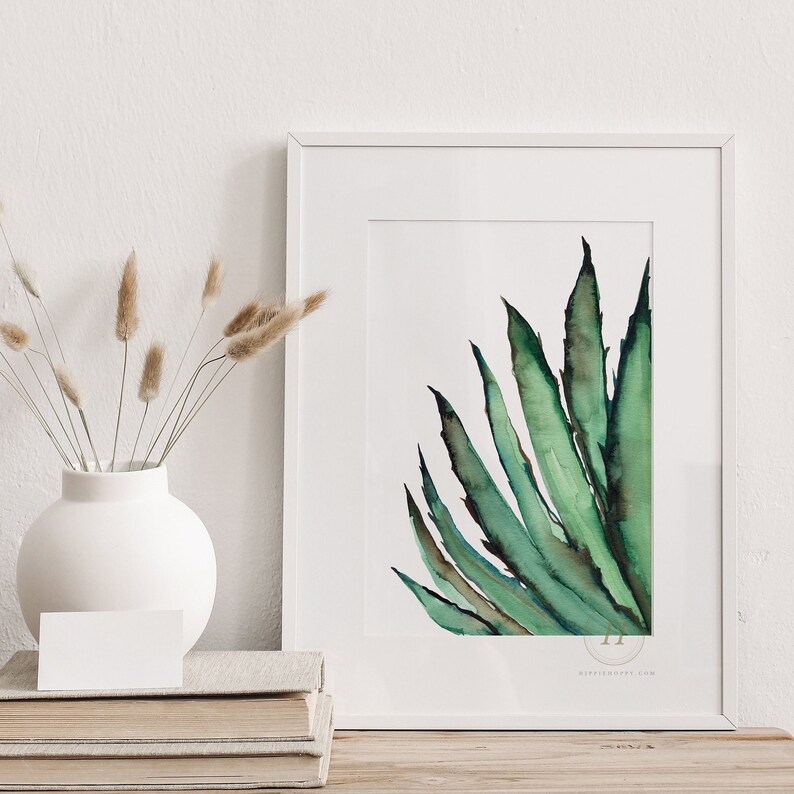 May include: A framed watercolor print of a green agave plant. The artwork features shades of green and brown, with a white border and frame. The print is displayed on a wooden surface, next to a white vase with dried flowers and a stack of books.