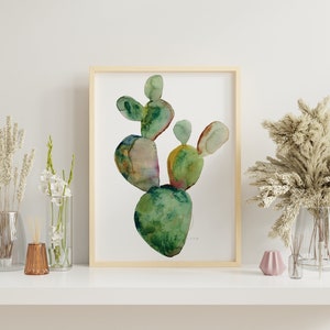 May include: A watercolor painting of a green cactus plant in a wooden frame. The cactus is painted in shades of green, yellow, and brown. The painting is on a white background.