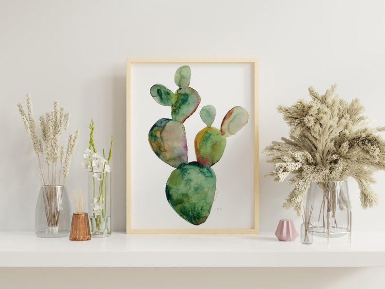May include: A watercolor painting of a green cactus plant in a wooden frame. The cactus is painted in shades of green, yellow, and brown. The painting is on a white background.