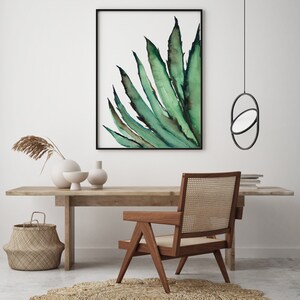 May include: A framed watercolor print of a green agave plant hangs above a wooden desk. A wooden chair with a woven back and a round jute rug are in front of the desk. A woven basket and decorative vases are also on the desk.