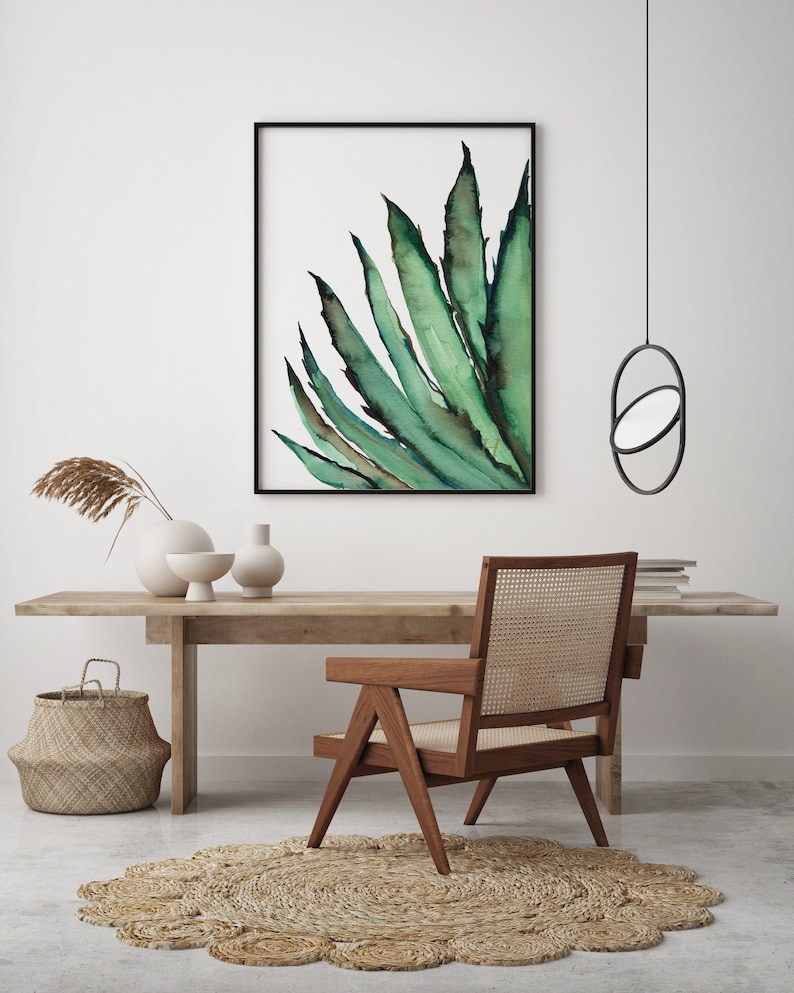 May include: A framed watercolor print of a green agave plant hangs above a wooden desk. A wooden chair with a woven back and a round jute rug are in front of the desk. A woven basket and decorative vases are also on the desk.