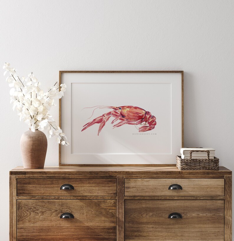 May include: A watercolor painting of a red crawfish in a wooden frame. The painting is on a wooden dresser with a vase of white flowers.
