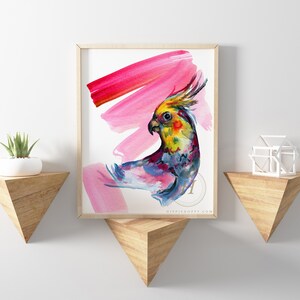 May include: A framed watercolour print featuring a vibrant cockatiel with a yellow crest, set against pink brushstrokes. The artwork is displayed on a white wall, with wooden shelves holding a plant and a geometric sculpture.