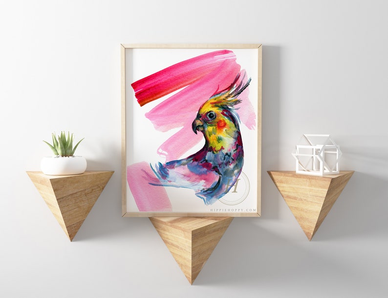 May include: A framed watercolour print featuring a vibrant cockatiel with a yellow crest, set against pink brushstrokes. The artwork is displayed on a white wall, with wooden shelves holding a plant and a geometric sculpture.