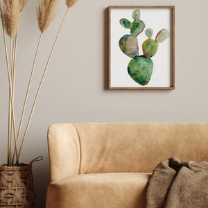 May include: A framed watercolor painting of a green cactus plant with a white background. The cactus is painted in a stylized, abstract style with shades of green, yellow, and brown.