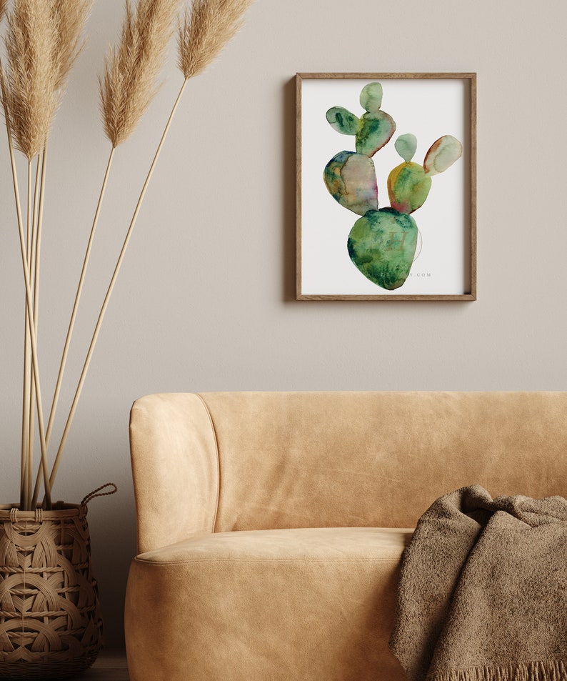 May include: A framed watercolor painting of a green cactus plant with a white background. The cactus is painted in a stylized, abstract style with shades of green, yellow, and brown.