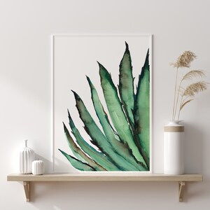 May include: A framed watercolor print of a green agave plant. The artwork features long, pointed leaves in shades of green and brown against a white background. The print is displayed on a light wood shelf with white vases and decorative dried plants.