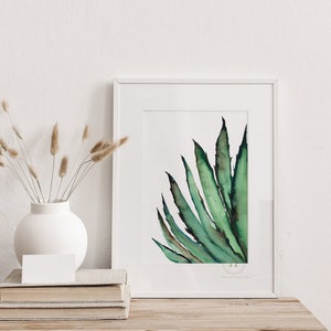 May include: A framed watercolor painting of a green agave plant. The artwork is displayed on a wooden table, next to a white vase with dried plants and a stack of books. The painting has a white border.