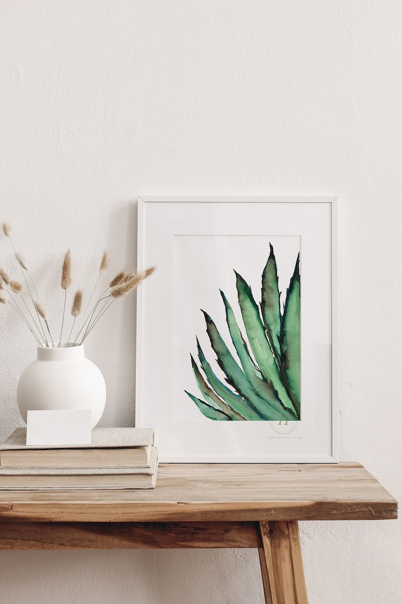 May include: A framed watercolor painting of a green agave plant. The artwork is displayed on a wooden table, next to a white vase with dried plants and a stack of books. The painting has a white border.