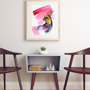 May include: Watercolour painting of a colourful cockatiel with a pink background, framed in a light wood frame. The artwork hangs above a small, blue side table with wooden legs and a small succulent plant.