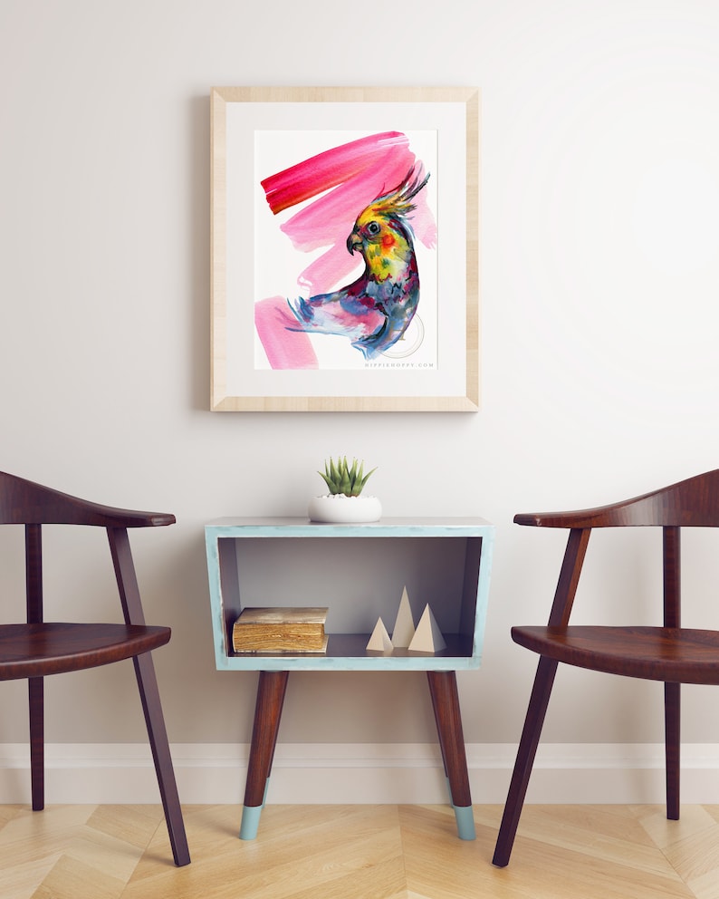 May include: Watercolour painting of a colourful cockatiel with a pink background, framed in a light wood frame. The artwork hangs above a small, blue side table with wooden legs and a small succulent plant.