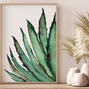 May include: Watercolor painting of a green agave plant with long, pointed leaves. The artwork is framed in a light wood frame and displayed on a wooden surface. Decorative vases and dried plants are in the background, creating a natural, botanical theme.