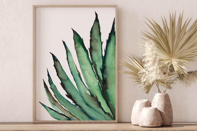 May include: Watercolor painting of a green agave plant with long, pointed leaves. The artwork is framed in a light wood frame and displayed on a wooden surface. Decorative vases and dried plants are in the background, creating a natural, botanical theme.
