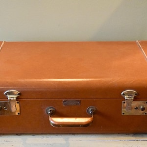 May include: A vintage brown suitcase with brass hardware, including latches, a handle, and corner protectors. The suitcase has a rectangular shape and is stitched with white thread along the edges. The suitcase is a classic travel accessory.