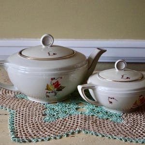 May include: A white teapot and sugar bowl with a floral design in red, yellow, and green. The teapot has a gold trim around the lid and the sugar bowl has a gold trim around the rim.