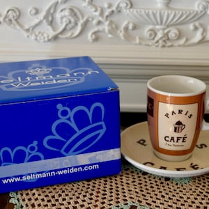 Peut inclure: Une boîte bleue avec une couronne blanche et le texte "Seltmann Weiden" et "www.seltmann-weiden.com". Une tasse à café blanche avec un motif brun et or qui dit "Paris Cafe" est sur une soucoupe blanche.