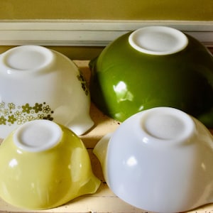 May include: Four vintage Pyrex mixing bowls in white, green, and yellow. One white bowl has a floral pattern in green. The bowls have a rounded shape with a handle on each side. The bowls are arranged on a wooden surface.