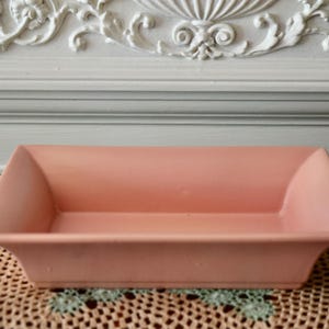 May include: A rectangular, light pink ceramic planter with a slightly tapered design. The planter sits on a decorative, round, crocheted doily with a scalloped edge. The background features a white, ornate wall molding.