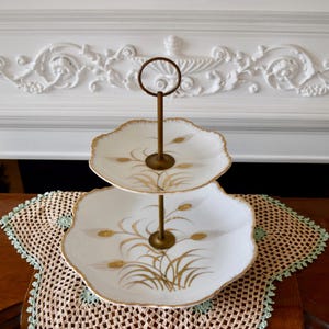 Peut inclure: Un support à gâteau en porcelaine blanche à deux étages avec une bordure dorée et un motif floral délicat. Le support a une poignée en métal et est parfait pour servir des desserts ou des en-cas.